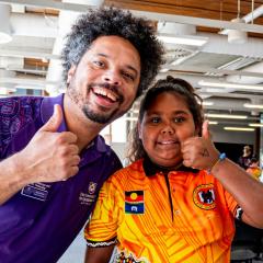Young girl in orange shirt and UQ staff member in purple shirt looking directly at the camera and giving thumbs up